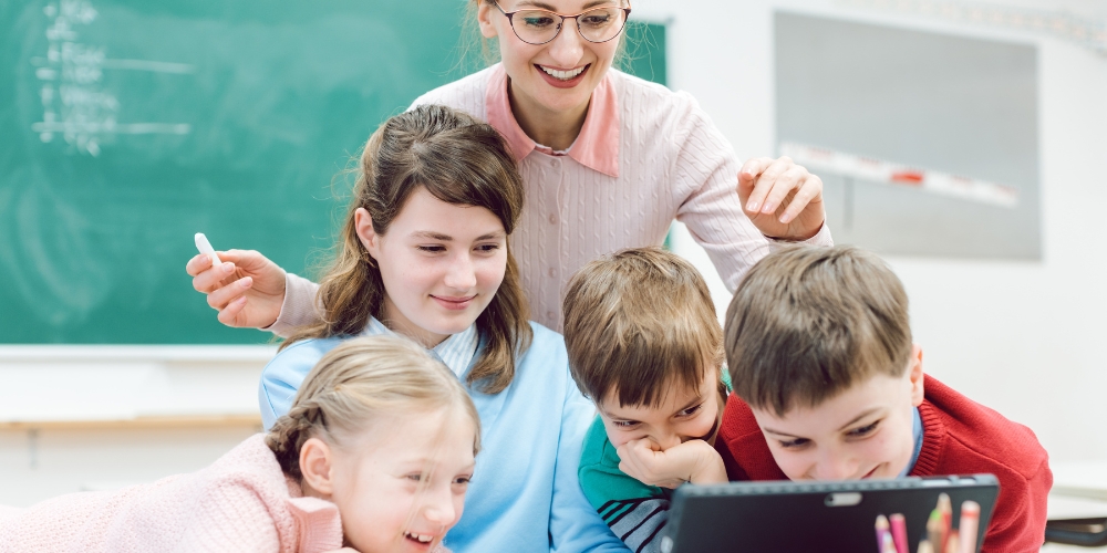 teacher standing behind four middle grade students looking at a tablet with a blackboard in background